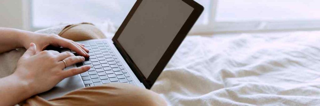 A person on a bed with white linen sheets. They are using their laptop which is sitting in their lap. 